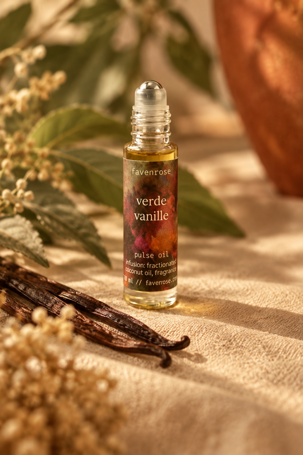 A small glass rollerball bottle labeled 'favenrose verde vanille' sits on a beige fabric surface with dried vanilla beans, green leaves, and soft warm lighting in the background.